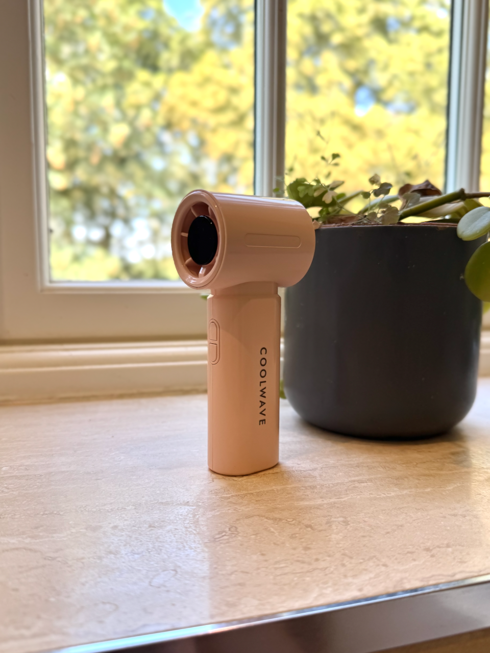 Pink CoolWave handheld fan standing beside a plant in a sunlit window